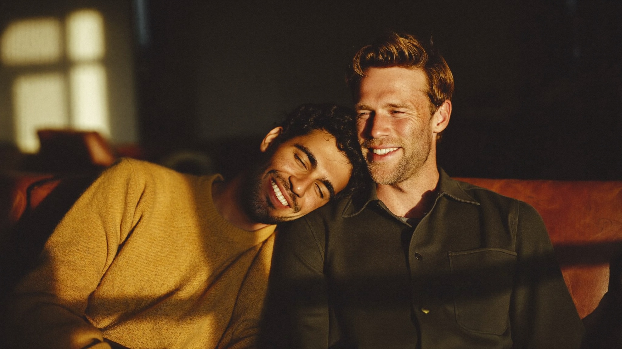 Two men laughing together on a sofa in golden afternoon light, one resting his head on the other's shoulder